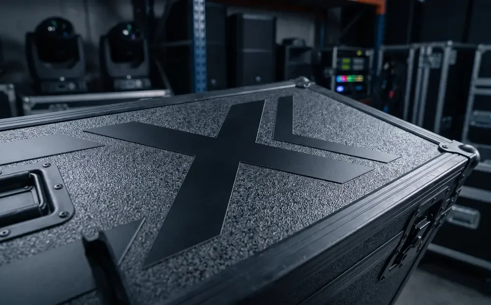 Schwarzes Transportcase mit X-Logo im Technik-Lager, dahinter Moving Heads und Lautsprecher im Regal