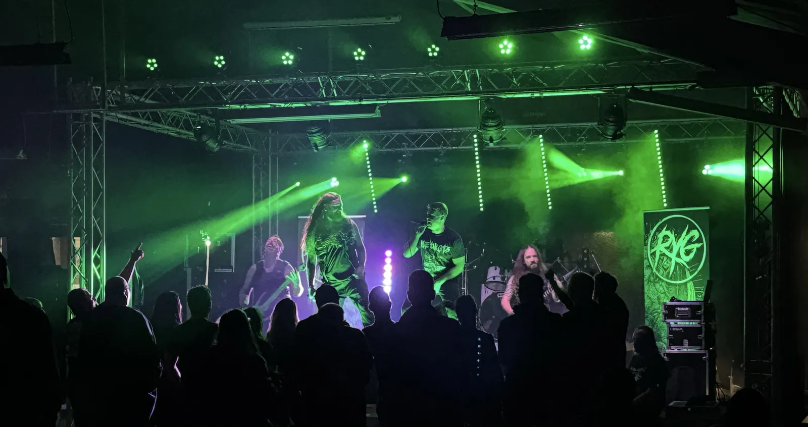 Metalband auf der Hausrock-Bühne mit Traversen in grünem Licht, Publikum als Silhouetten davor